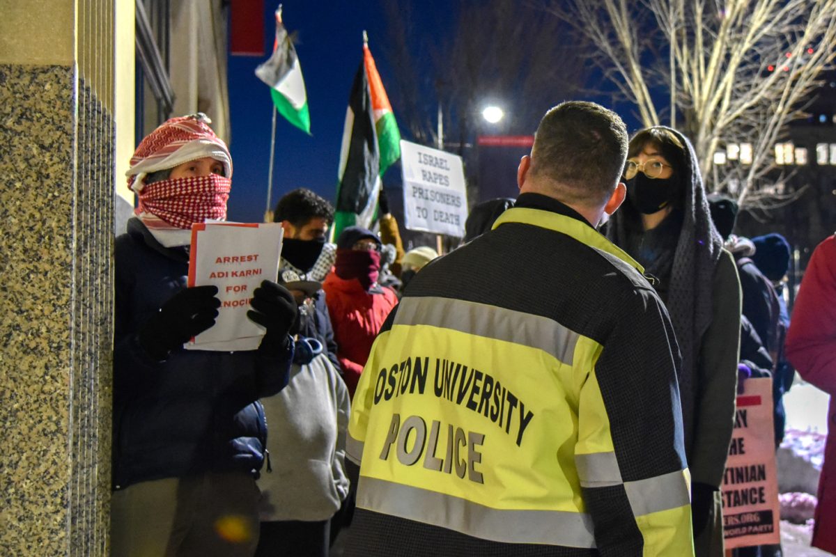 A masked protester outside the Howard Thurman Center for Common Ground holds up a sign reading “ARREST ADI KARNI FOR GENOCIDE.” 