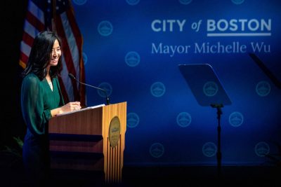 Mayor Wu standing and smiling behind a podium. "City of Boston Mayor Michelle Wu" is projected behind her