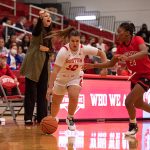 Women's basketball player dribbles away from an opponent while coach watches