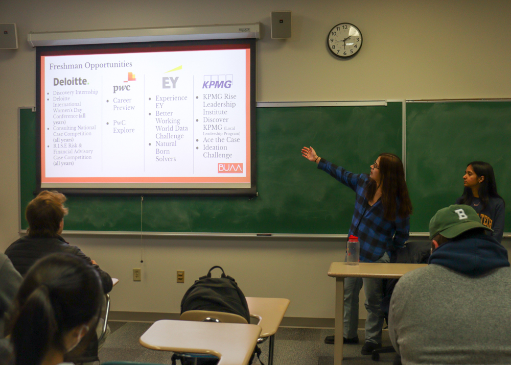 A person pointing toward a screen and speaking to a classroom of people.