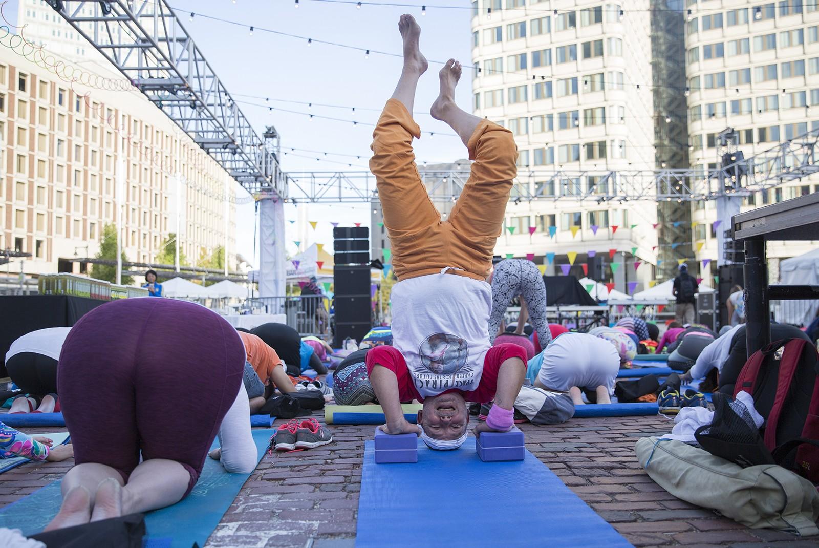 Gary K. Wolf, 75, of Brookline, performs a Sirsasana headstand while other participants perform Bālāsana, or Child’s Pose, at “Daybreaker BOS presents: 80s Throwback - Jazzercise Edition” in City Hall Plaza early Saturday morning. PHOTO BY SARAH SILBIGER/ DAILY FREE PRESS STAFF