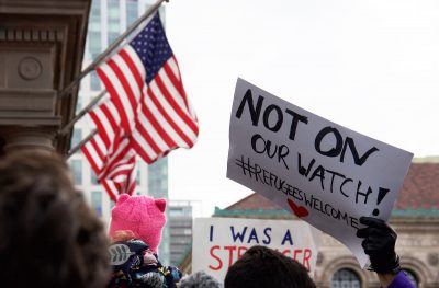 A protester holds sign in Copley Square while a U.S. flag waves in the background. PHOTO BY OLIVIA FALCIGNO/ DAILY FREE PRESS STAFF